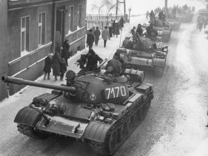A city street with a line of tanks driving down the road in the snow and people walking on the sidewalks