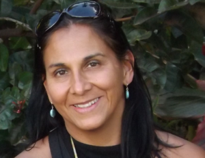 Carmen smiling and standing in front of greenery, wearing sunglasses on their head, long light blue earrings and a black high necked shirt. 