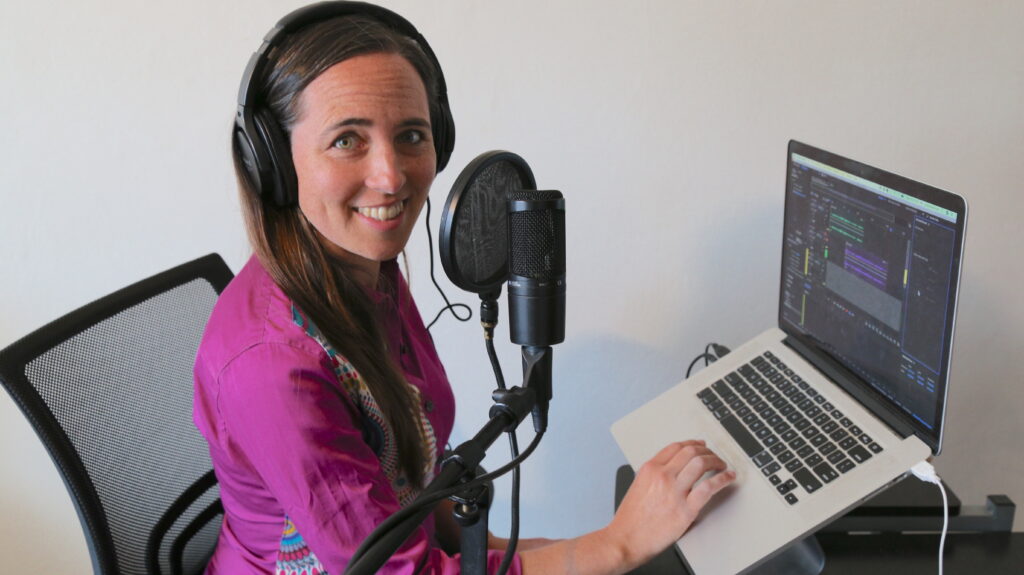 Sarah Van Borek sits with one hand on the keyboard of her laptop, turning and smiling at the camera. A larger microphone sits between the viewer and Sarah, who sports large headphones. 