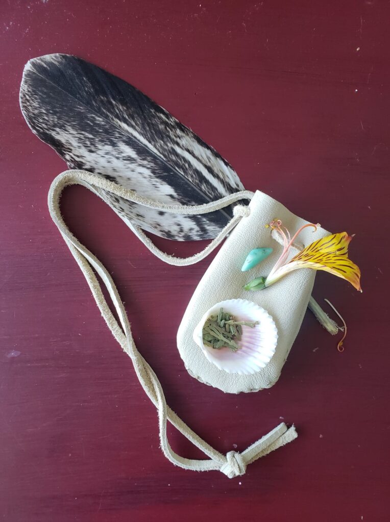 A white leather medicine bag with a turquise stone, yellow flower, and black and white feather sits on a dark purple table