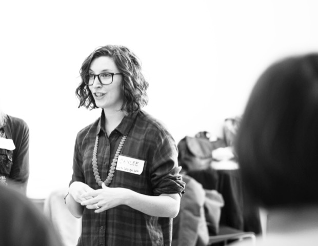 Caylee, sporting glasses, a plaid shirt and name tag, speaks to a group of people