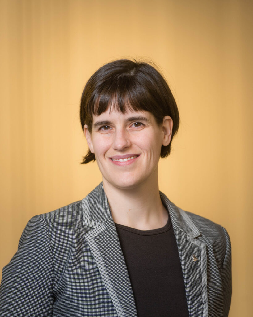 Agnes wearing a grey blazer and black shirt, smiling in front of a yellow background