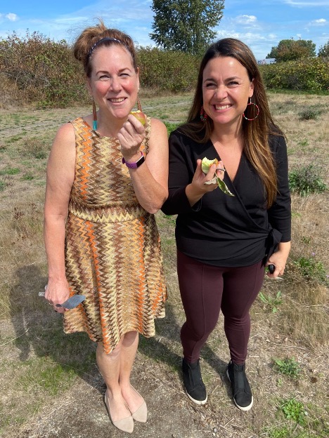 Valerie and Katie stand side by side, smiling and eating apples