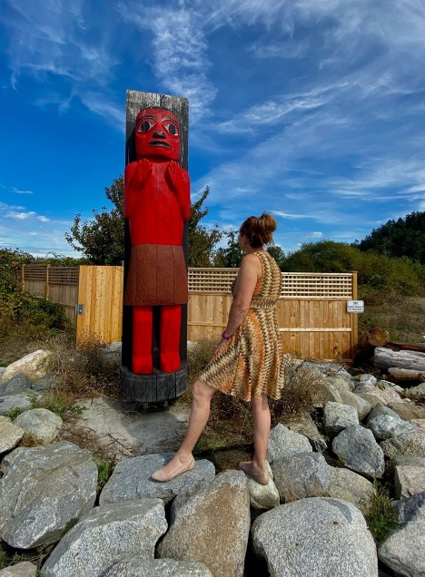 Valerie is turned to face a large red wooden carving known as a house post (similar to a totem pole).