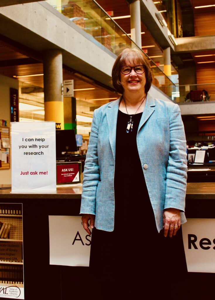 Linda stands in a library. A sign next to her says "I can help you with your research. Just ask me!"