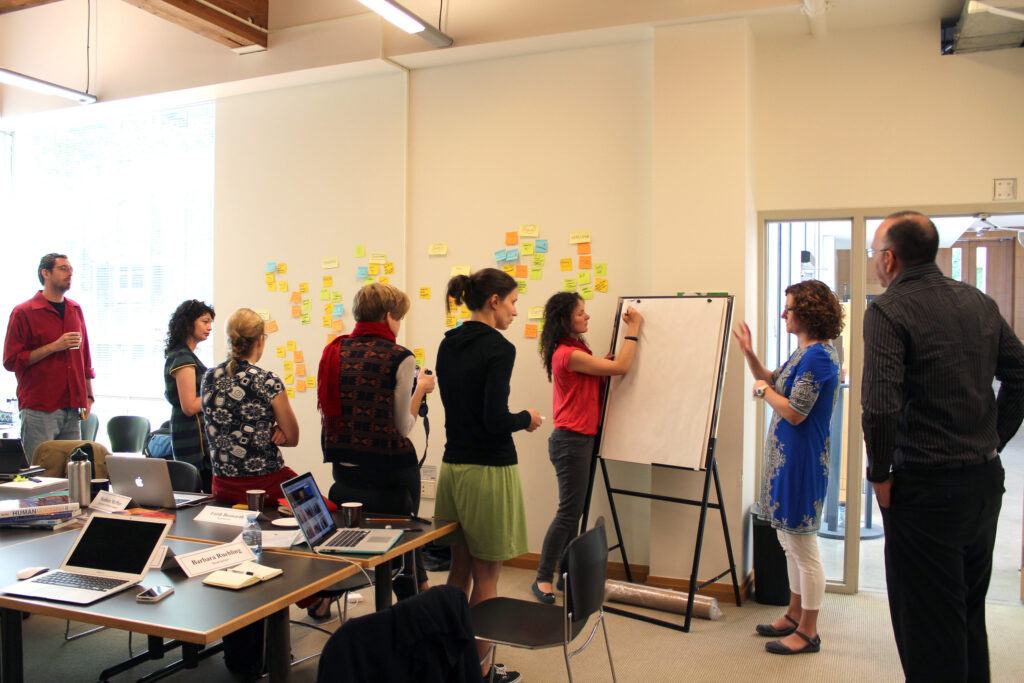 a group of 8 people brainstorm in front of white board, many sticky notes are seen in the background