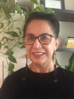 Debra Hoggan smiles in an office setting. She wears dark rimmed glasses and beaded earrings that graze her black shirt.