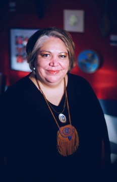 Inez Cook in what appears to be an art gallery. She is smiling and wearing a long black sweater with a necklace that looks like a leather medicine bag.