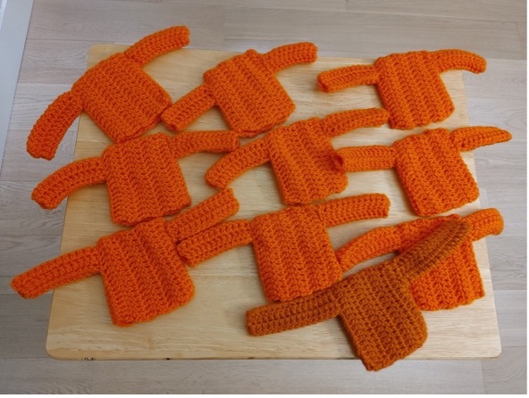 10 tiny crocheted orange shirts sit on a wooden surface