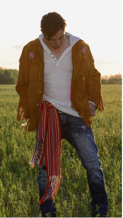 Stephen Gladue stands in a field of green grass. He is wearing a white shirt, jeans, a brown jacket with beaded embellishments and a Metis sash is tied at this waist.