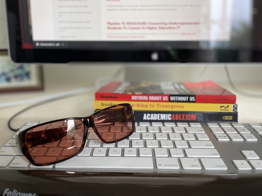 pink tinted glasses sit on a computer keyboard. Three books are stacked in the background: "Nothing About As Without Us", "Teaching to Transgress" and "Academic Ableism". 