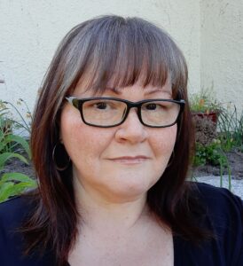 A middle-aged Indigenous woman with shoulder-length brown hair, bangs, and black rimmed glasses.