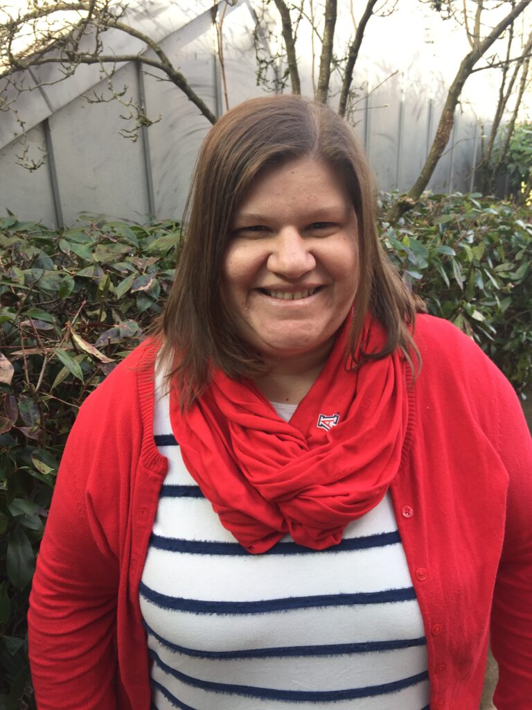 Middle-aged white woman with brown hair wearing a blue-and-white striped shirt, a red cardigan, and a scarf stands in front of a hedge.