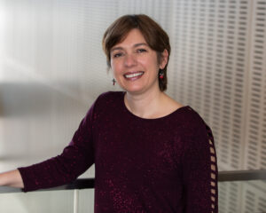 Adina Gray smiling in a burgundy top, standing indoors by a railing.