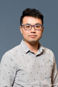 A medium close-up portrait of an Asian male against a solid gray-blue background. He is wearing dark, blue-rimmed glasses and a gray collared shirt with subtle dark feathered patterns. The man is looking directly at the camera with a small grin. The lighting is even, illuminating their face clearly.