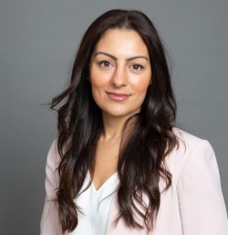 Professional headshot of Harroop Sharda, a woman with long wavy dark brown hair, wearing a light pink blazer over a white blouse, posing in front of a neutral gray background. She has a warm expression and is looking directly at the camera
