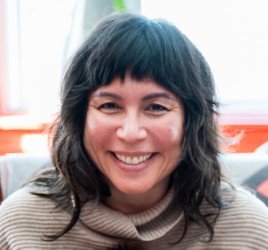 Allison is a mixed-race, Japanese American woman with long black hair and bangs wearing a beige, cowl-neck, textured sweater and smiling at the camera in a brightly lit, blurry interior space.