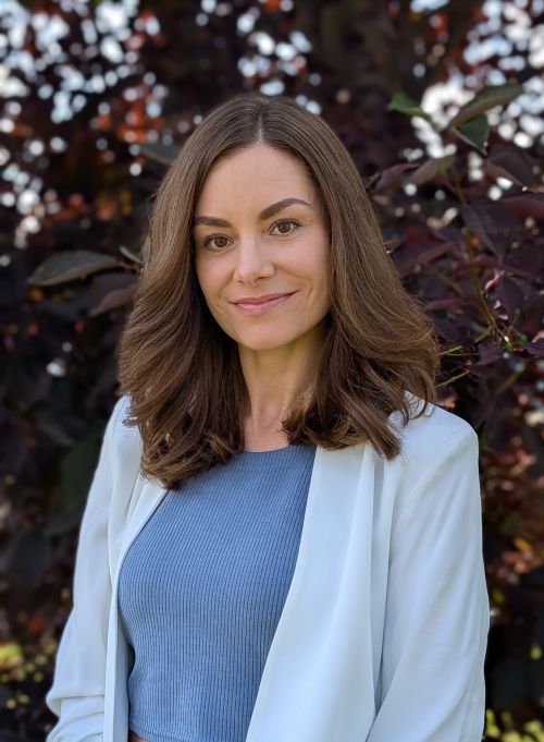 Darryn DiFrancesco: Headshot of a woman with shoulder-length brown hair wearing a light blue blazer over a blue top, photographed against a background of dark foliage 