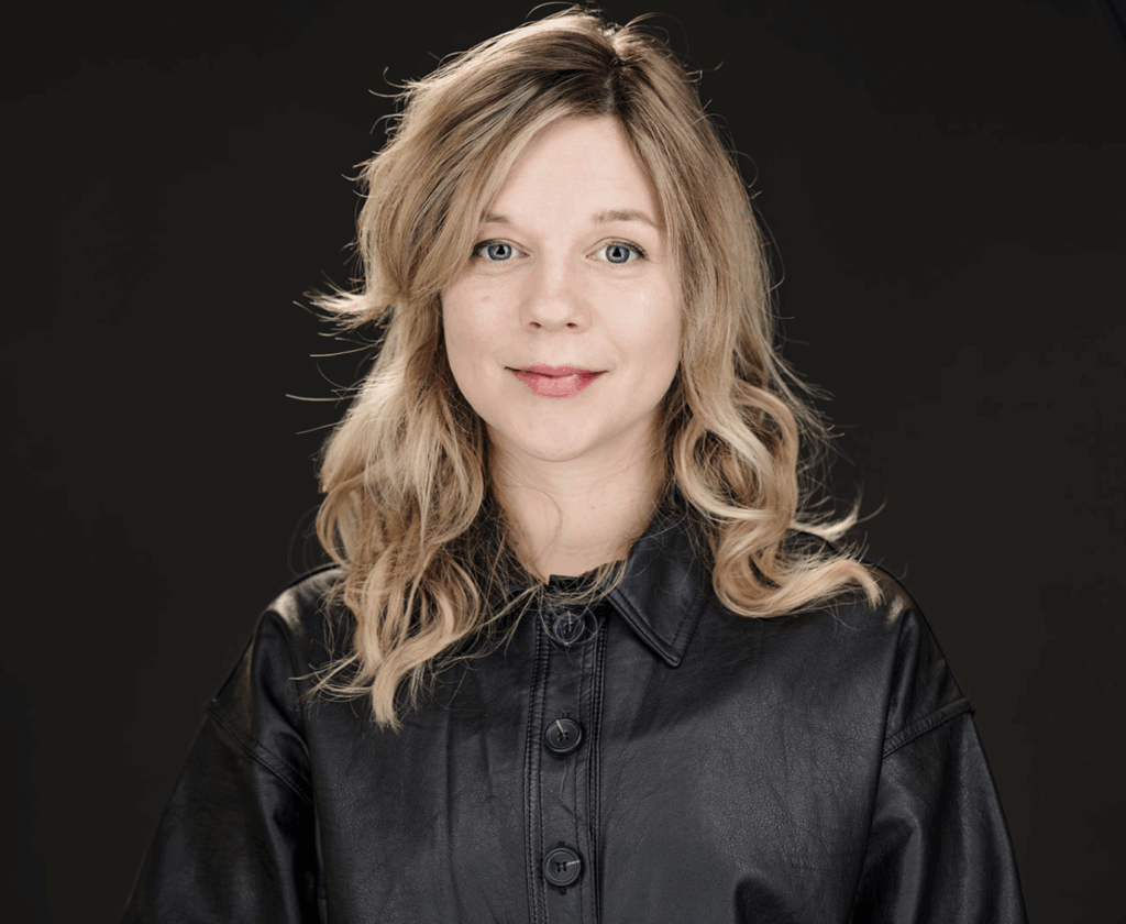 Sophia Jaworski: A blond femme person with shoulder length wavy hair smiling 